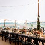 Elegant tented wedding reception with long tables, black chairs, candlelight, and colorful floral centerpieces overlooking countryside