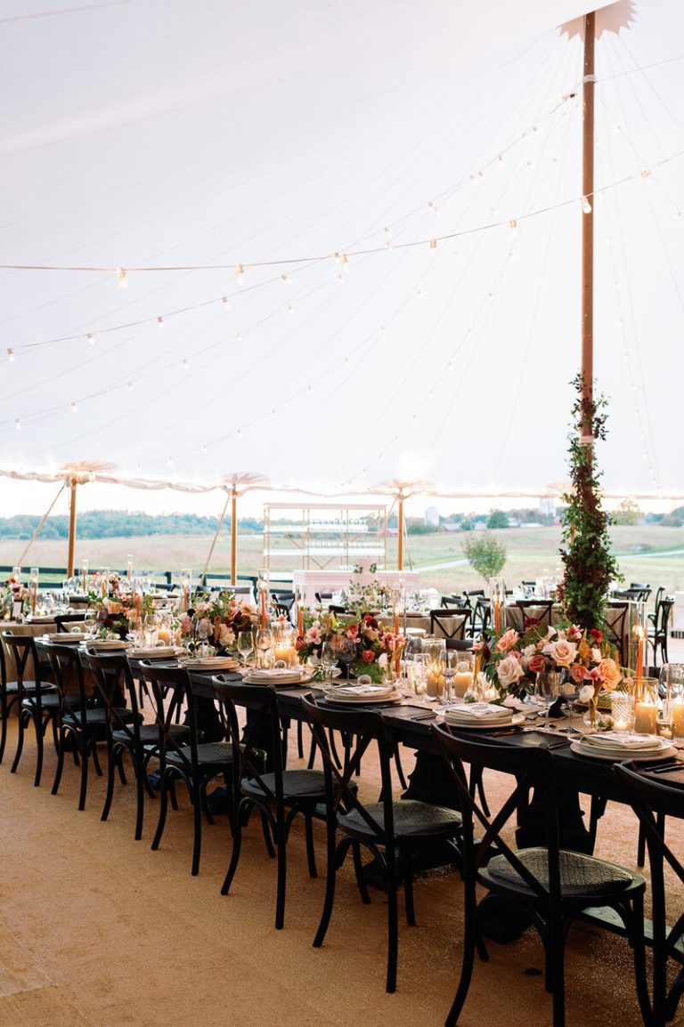 Elegant tented wedding reception with long tables, black chairs, candlelight, and colorful floral centerpieces overlooking countryside