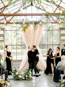Two grooms kissing under draped ceremony arch with greenery and string lights in glass greenhouse venue