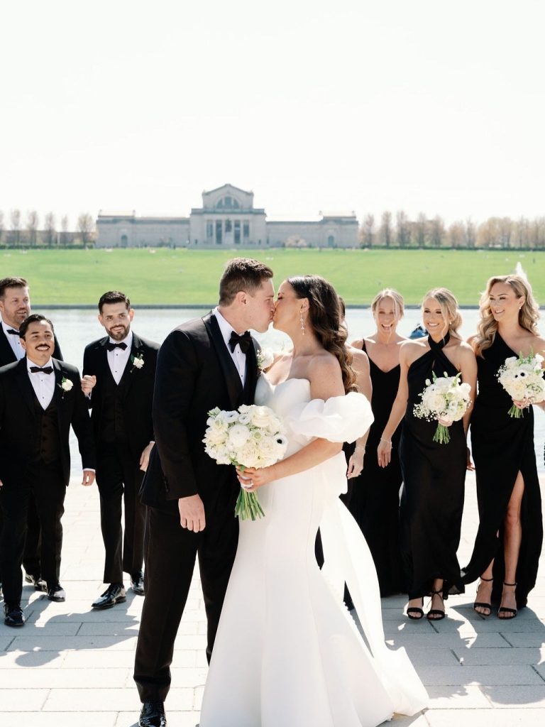 Bride and groom kissing surrounded by wedding party in black attire at St. Louis Art Museum