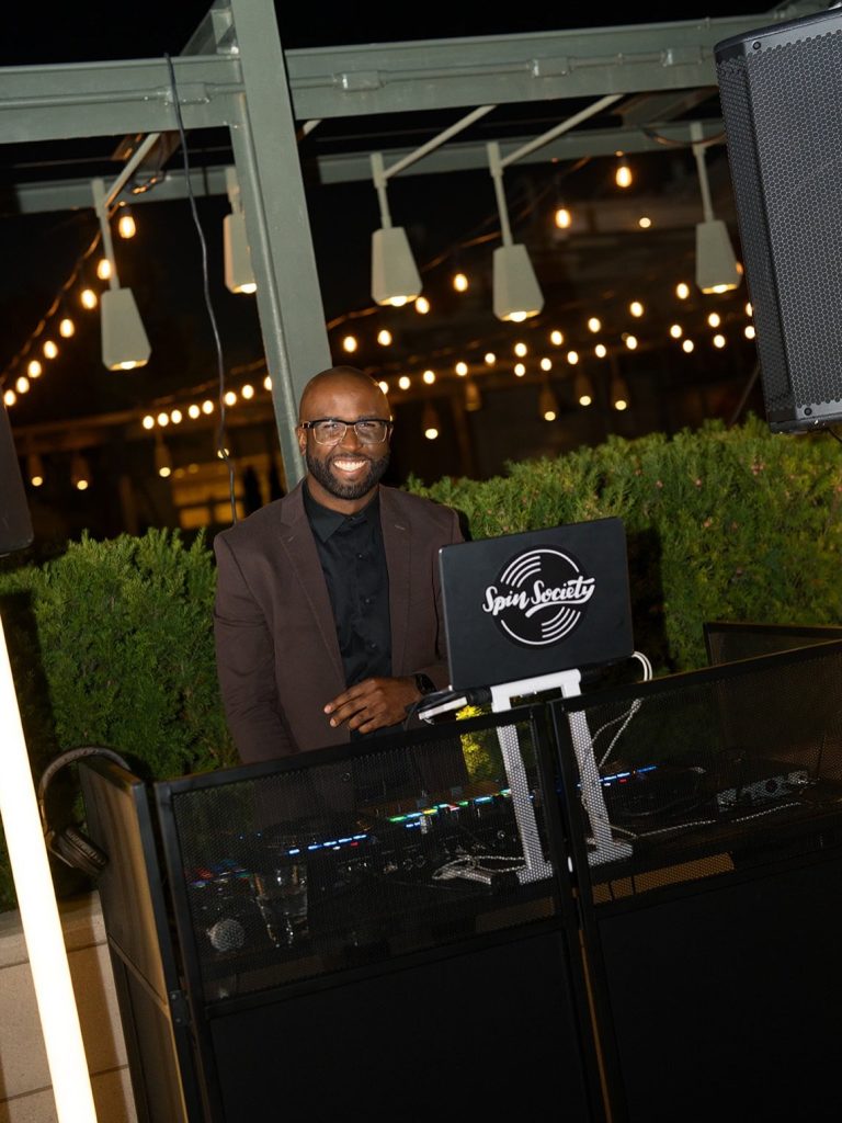 Spin Society DJ smiling behind his equipment setup at rooftop wedding venue with string lights