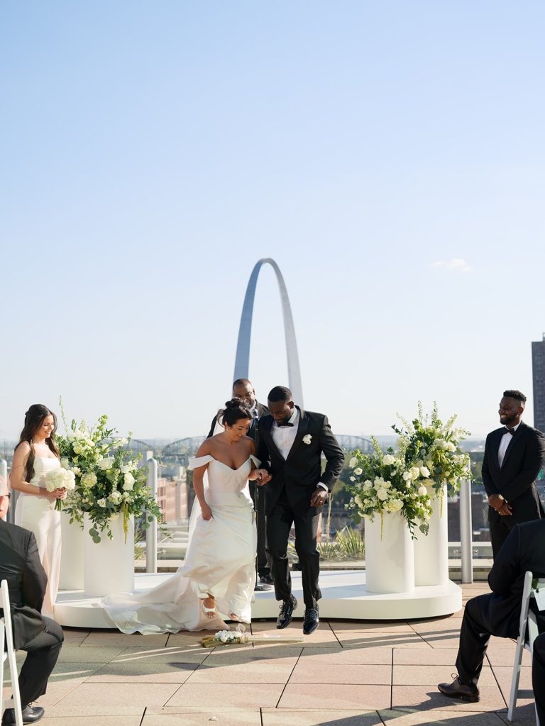 Katie and Anthony walking down the aisle after rooftop ceremony, Gateway Arch behind them, flanked by white floral arrangements