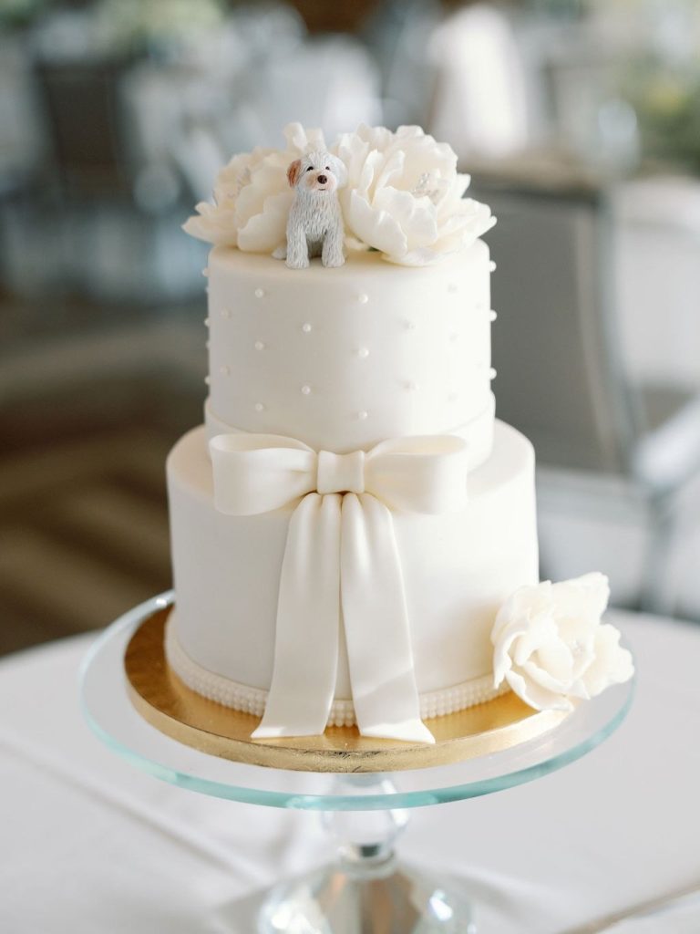 Two-tier white wedding cake with pearl dots, fondant bow, and dog figurine topper with sugar flowers