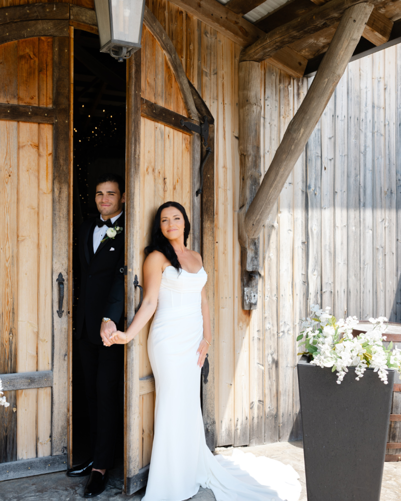 Bride and groom holding hands in rustic barn doorway with white floral arrangements