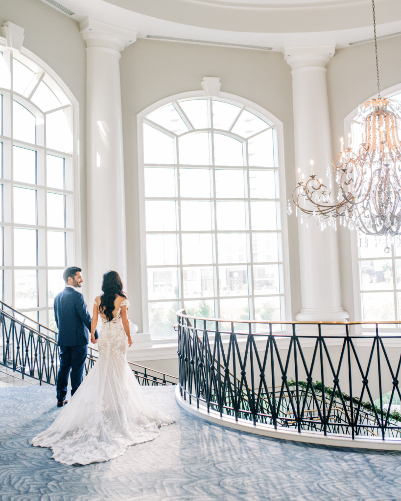 Bride and groom walk along curved balcony in elegant ballroom with arched windows and crystal chandelier