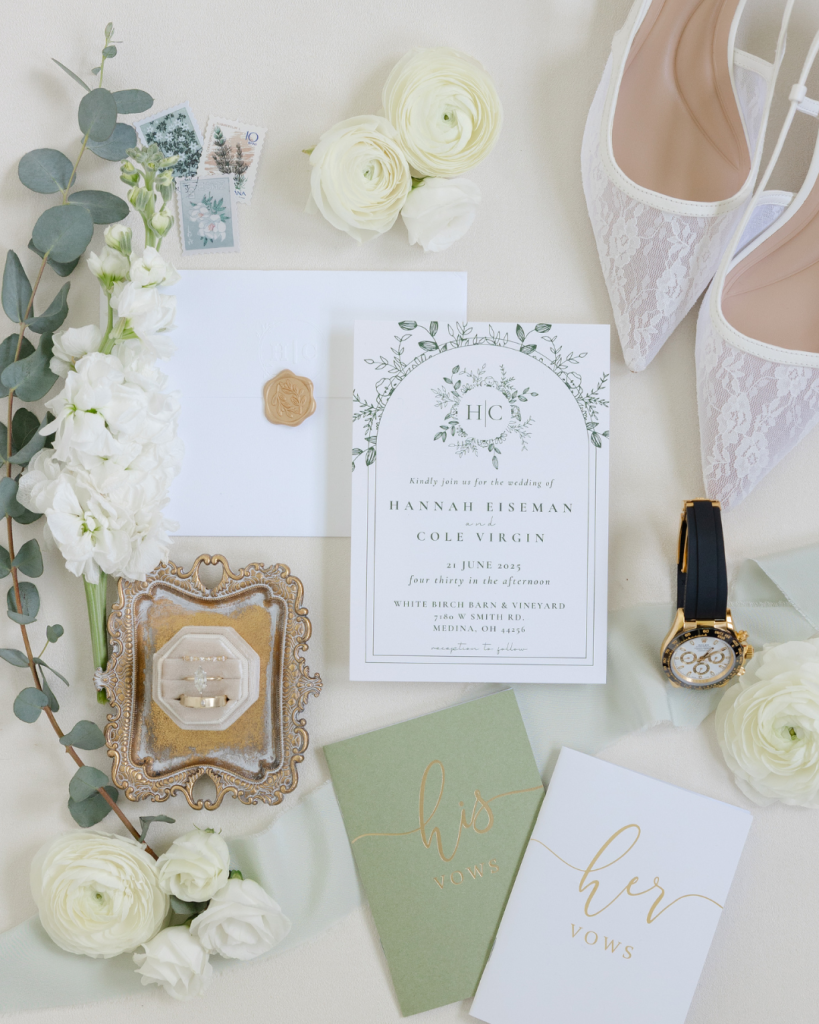Elegant wedding invitation suite with white ranunculus, eucalyptus, lace bridal shoes, and sage green vow books on ivory background