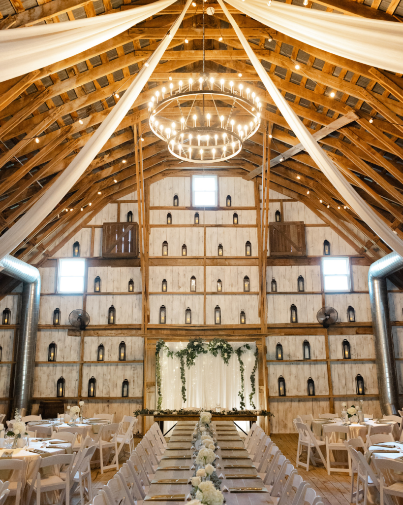 Rustic barn reception venue interior featuring wooden beams, circular chandelier, lantern-lined walls, and ceremony setup with white draping
