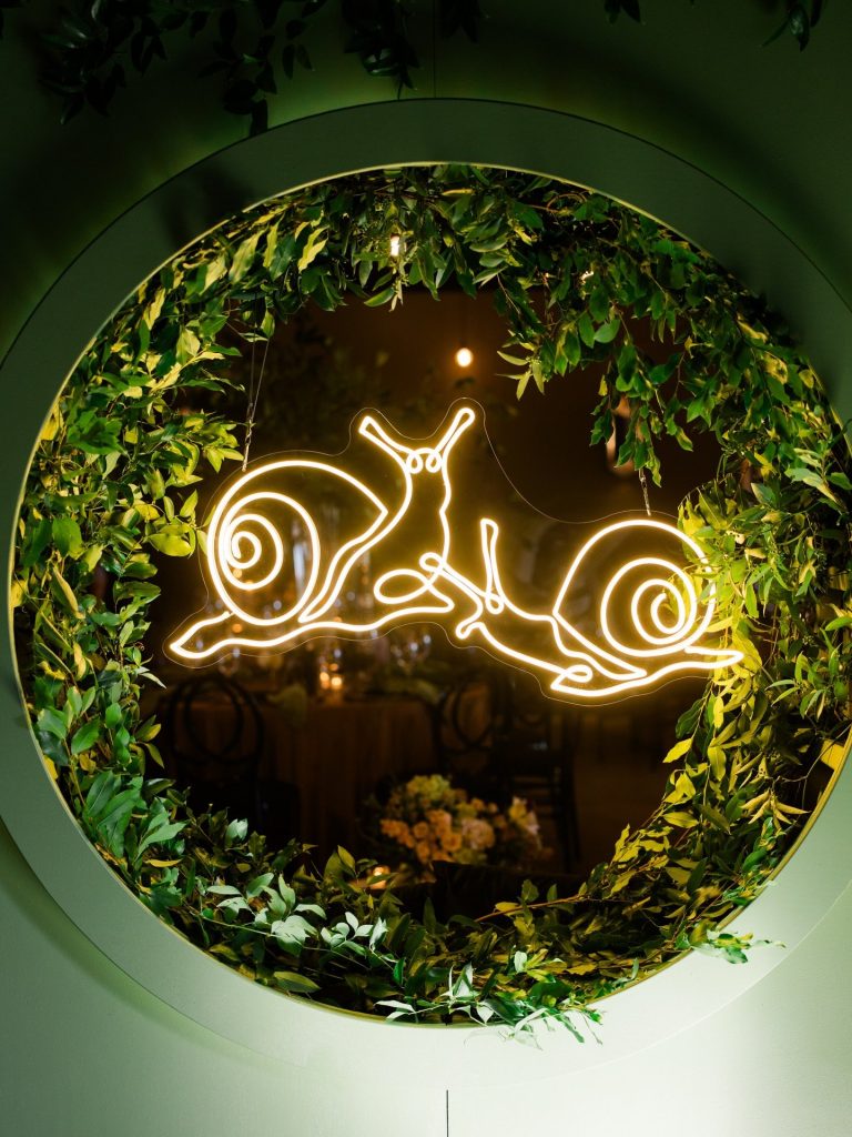 Illuminated neon snail sign framed by circular wreath of fresh greenery and warm lighting at wedding reception