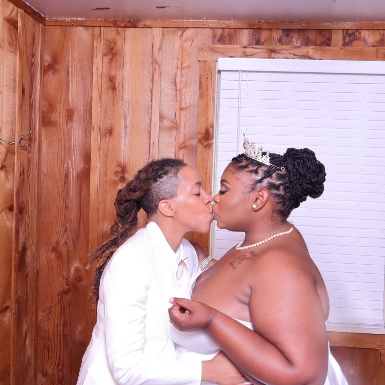 Two brides sharing a kiss in white formal wear against rustic wood paneling