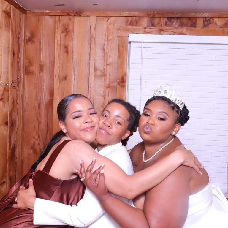 Three women embracing and smiling in a wedding photo booth, one wearing a tiara
