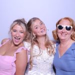Three women posing together in a photo booth, with the bride in a white dress flanked by bridesmaids in pink and blue