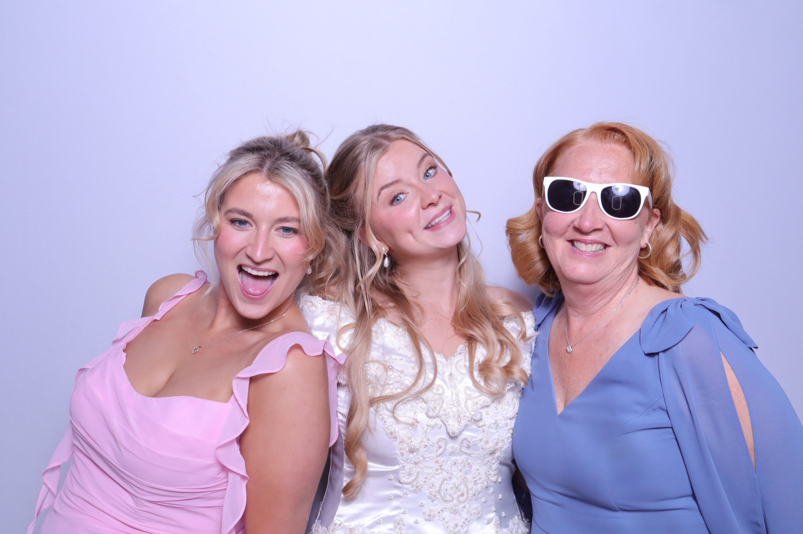 Three women posing together in a photo booth, with the bride in a white dress flanked by bridesmaids in pink and blue