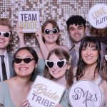 Wedding party holding "Team Bride," "Bride Tribe," "Love Him," and "Mrs" signs with heart-shaped sunglasses against sequin backdrop