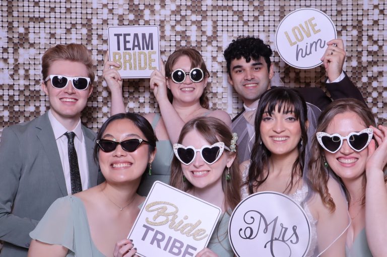 Wedding party holding "Team Bride," "Bride Tribe," "Love Him," and "Mrs" signs with heart-shaped sunglasses against sequin backdrop