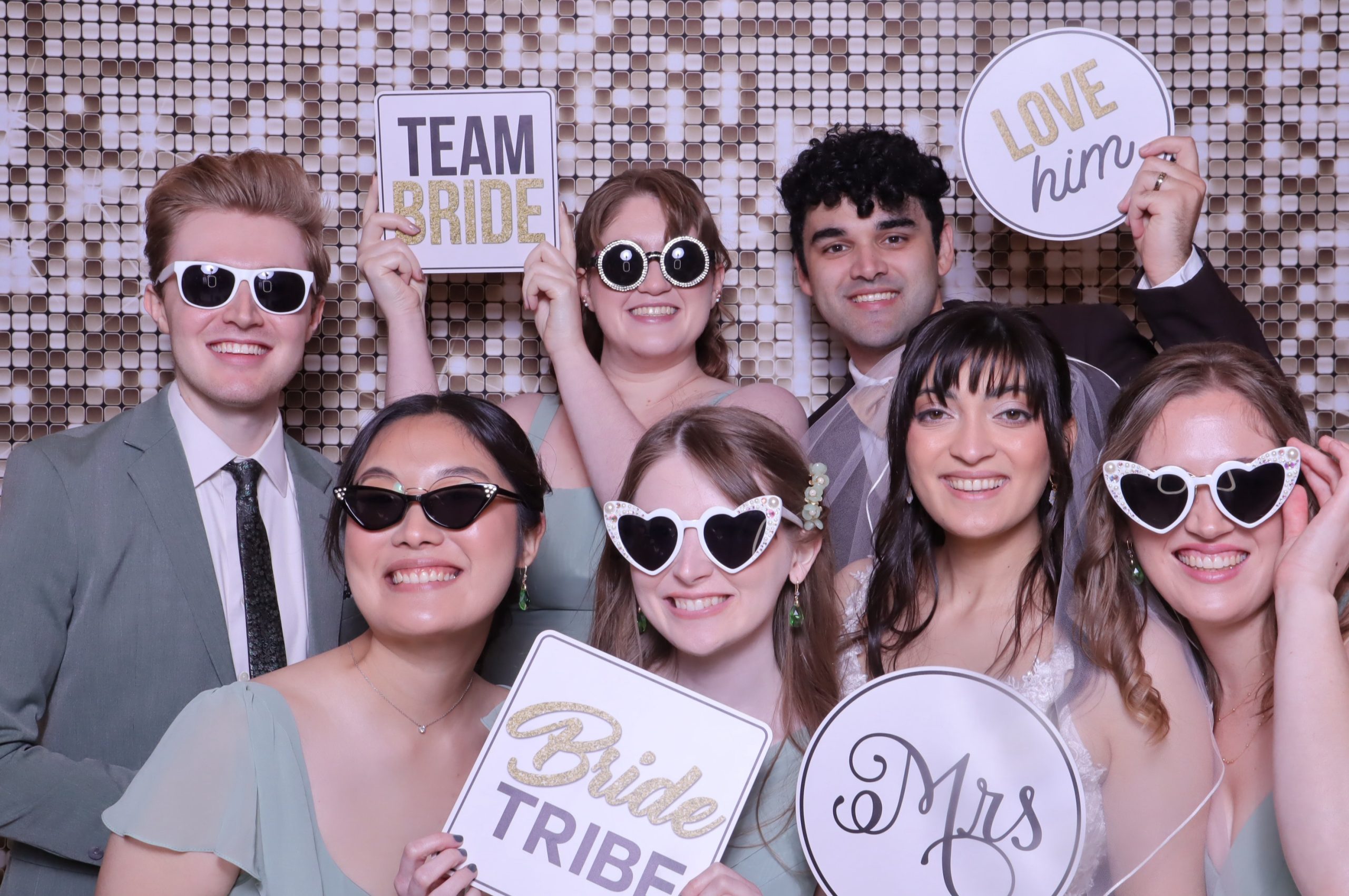 Wedding party holding "Team Bride," "Bride Tribe," "Love Him," and "Mrs" signs with heart-shaped sunglasses against sequin backdrop