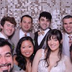 Bride and groom with wedding guests smiling in formal attire against metallic sequin wall