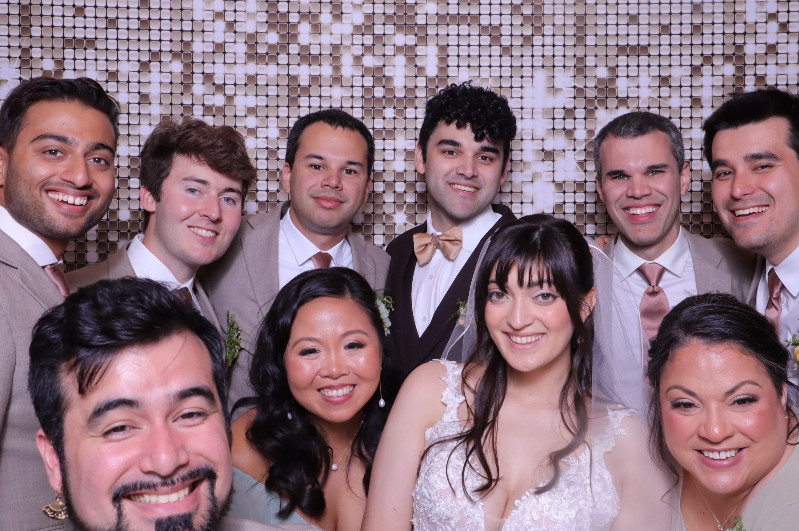 Bride and groom with wedding guests smiling in formal attire against metallic sequin wall