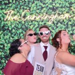 Wedding guests wearing novelty sunglasses posing with "Team Bride" sign against greenery wall backdrop with neon "The Cunningham's" sign
