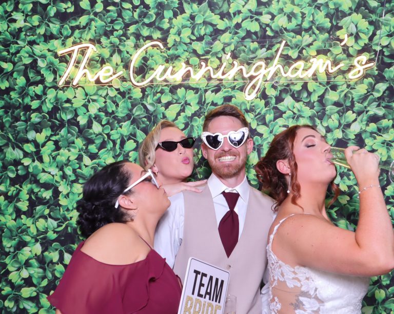 Wedding guests wearing novelty sunglasses posing with "Team Bride" sign against greenery wall backdrop with neon "The Cunningham's" sign