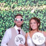 Bride and groom posing at wedding photo booth with Mr. and Mrs. signs against greenery wall backdrop with neon "The Cunningham's" sign