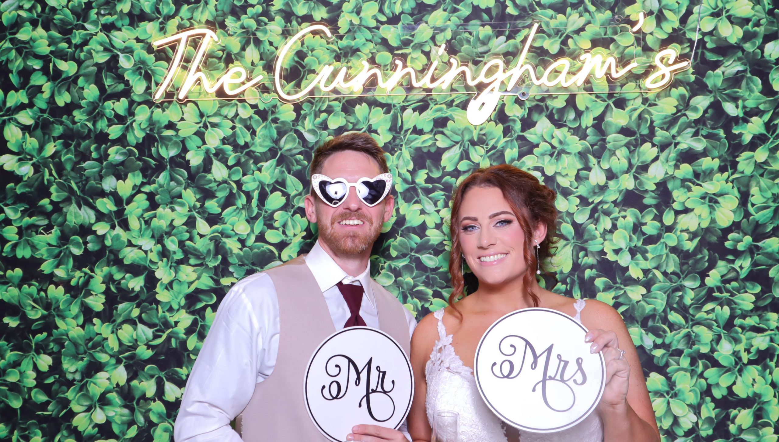 Bride and groom posing at wedding photo booth with Mr. and Mrs. signs against greenery wall backdrop with neon "The Cunningham's" sign