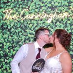 Bride and groom kissing in photo booth with greenery wall backdrop and custom neon sign reading "The Cunningham's"