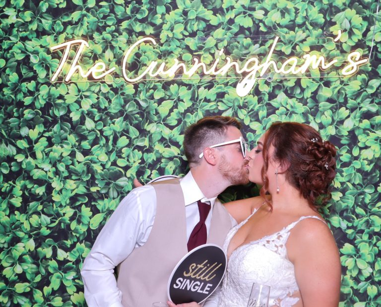 Bride and groom kissing in photo booth with greenery wall backdrop and custom neon sign reading "The Cunningham's"
