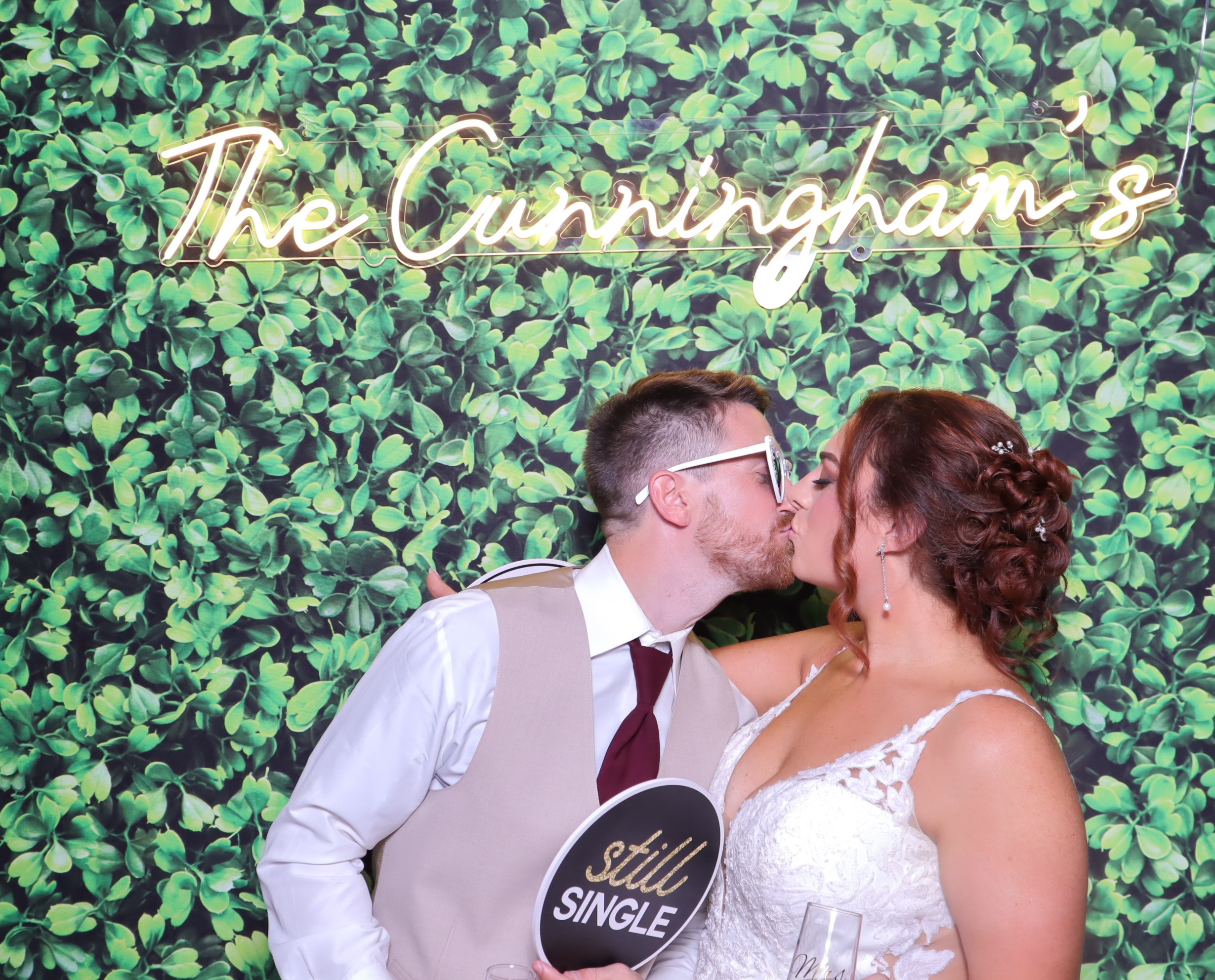 Bride and groom kissing in photo booth with greenery wall backdrop and custom neon sign reading "The Cunningham's"