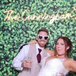 Bride and groom with champagne and heart sunglasses posing at greenery wall backdrop