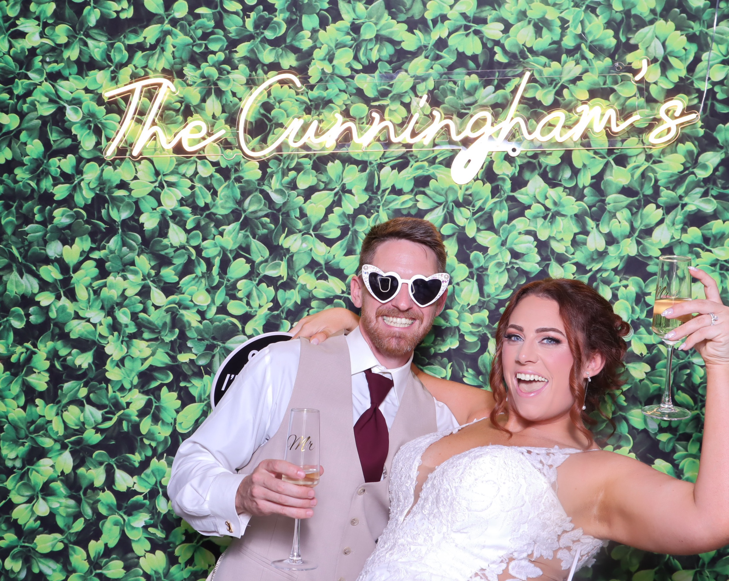 Bride and groom with champagne and heart sunglasses posing at greenery wall backdrop