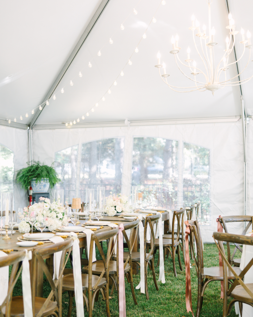 White tent reception space with wooden farm tables, cross-back chairs with pink ribbon accents, and string lights overhead