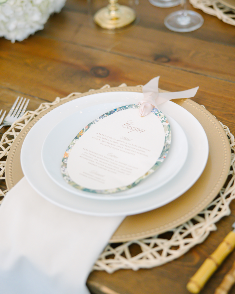 White dinner plate on gold charger with floral-bordered menu card and ribbon detail on wooden farmhouse table