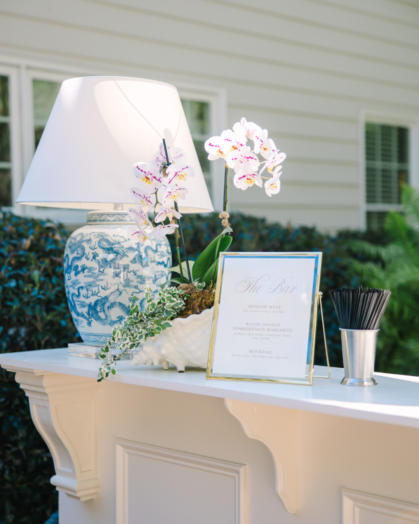 Elegant bar setup with blue and white chinoiserie lamp, white orchid, framed bar menu, and cocktail stirrers