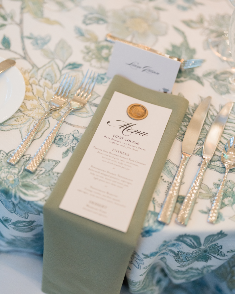 Sage green menu card with gold wax seal and pearl-handled silverware on botanical print linens