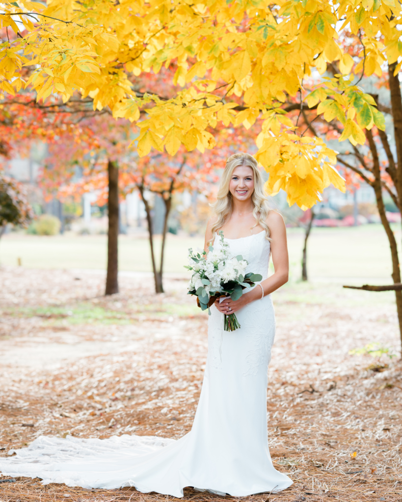 Bride in lace gown holding white and eucalyptus bouquet beneath vibrant golden yellow autumn trees