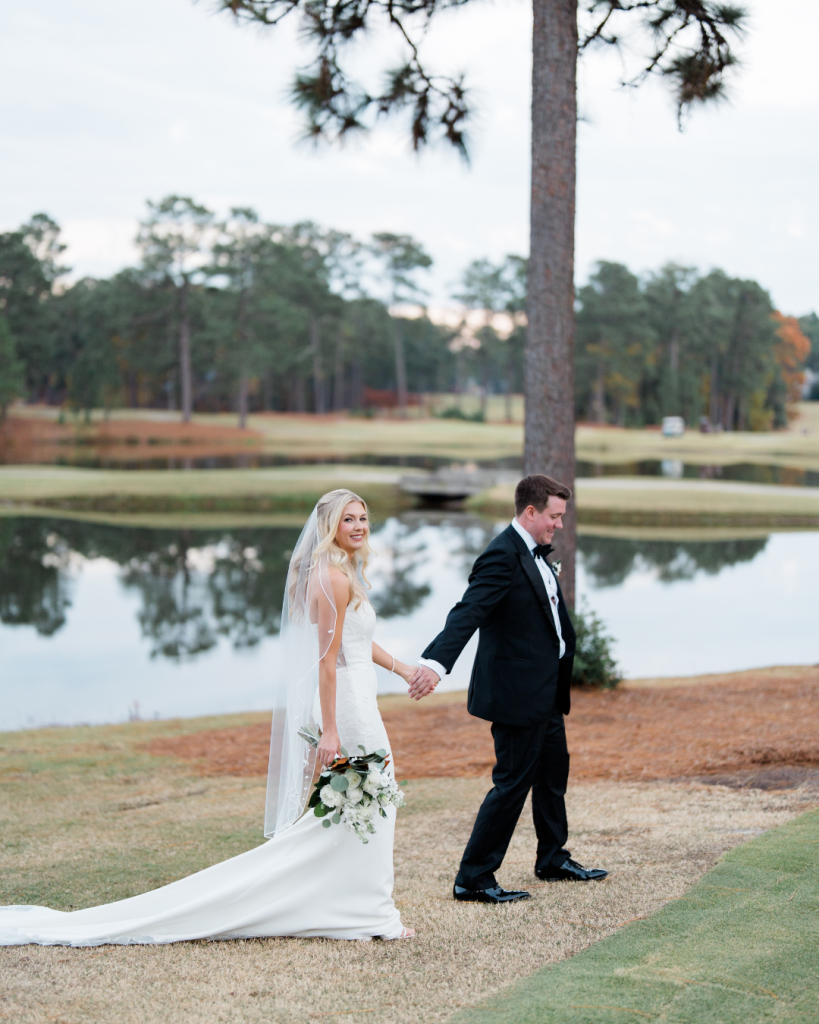 Bride and groom holding hands while walking along golf course pond at sunset