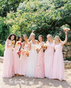 Bride in white gown with six bridesmaids in pink dresses holding colorful bouquets in garden setting