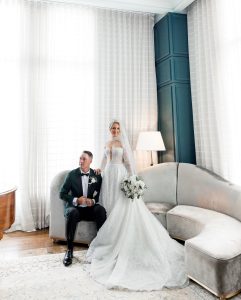 Bride in beaded long-sleeve gown with cathedral veil standing beside groom in dark green tuxedo in luxury hotel suite