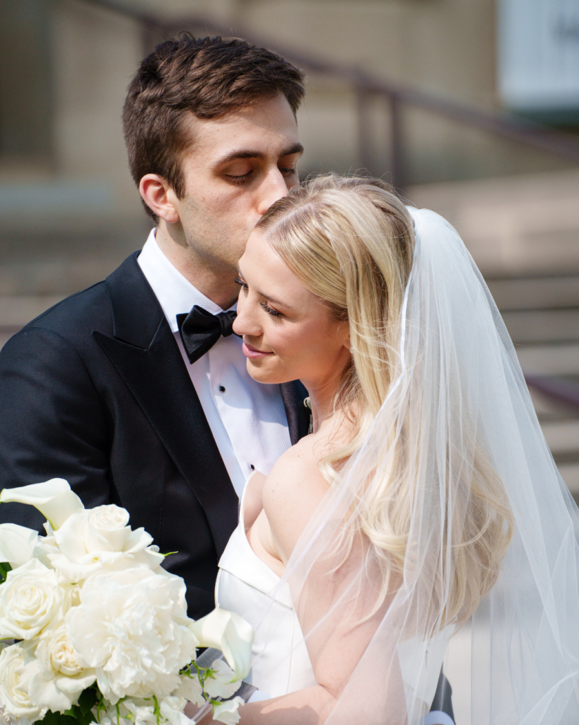 Kyle kisses Marielle's forehead as she holds white rose bouquet, veil flowing behind her