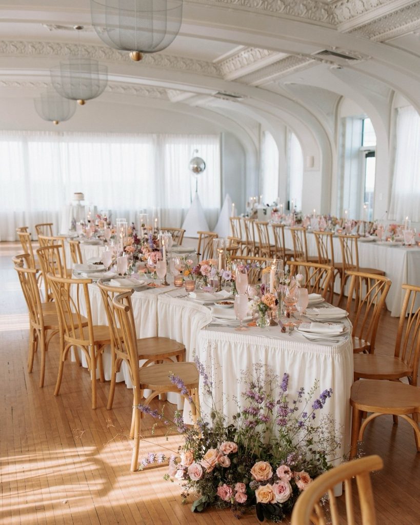 Elegant wedding reception hall with white ornate ceiling, long tables with pink floral centerpieces, and natural wood chairs