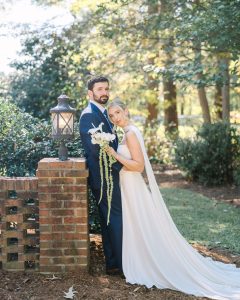 Couple in wedding attire embracing near brick pillar surrounded by lush greenery