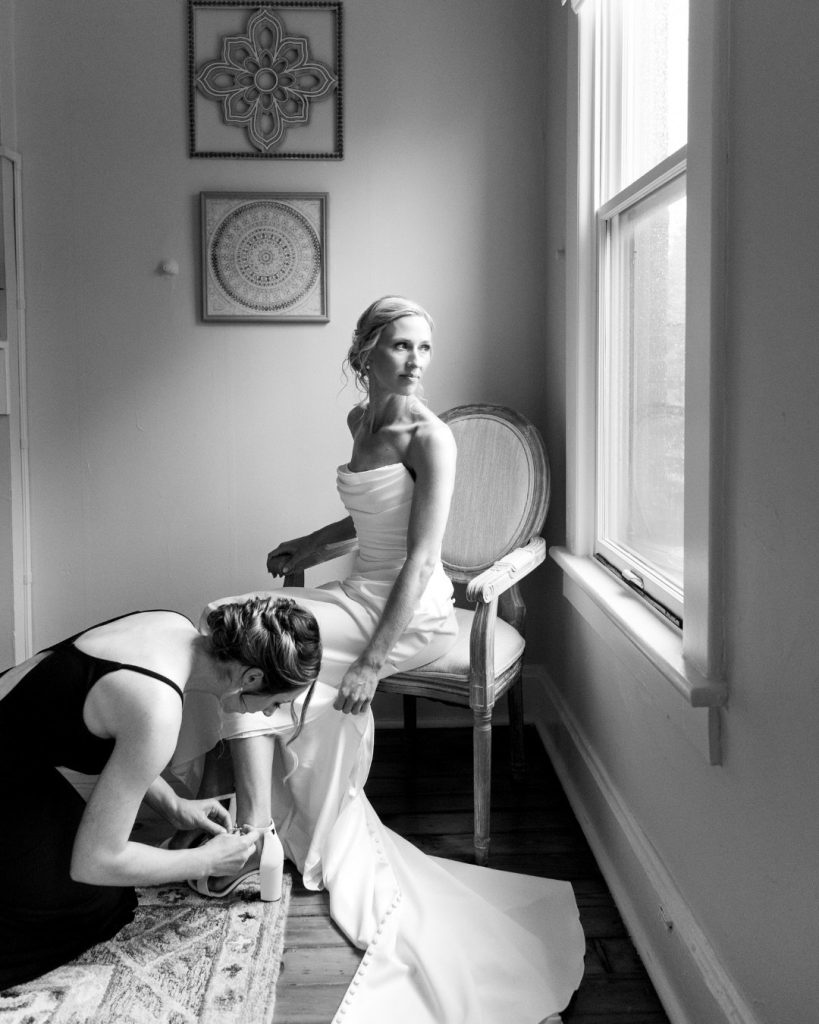 Bridesmaid fastens bride's wedding shoes as she sits in vintage chair by window