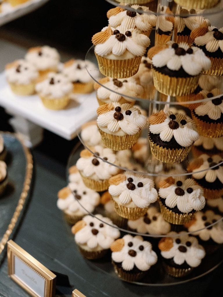 Tiered display of cupcakes with white and gold frosting topped with chocolate decorative accents