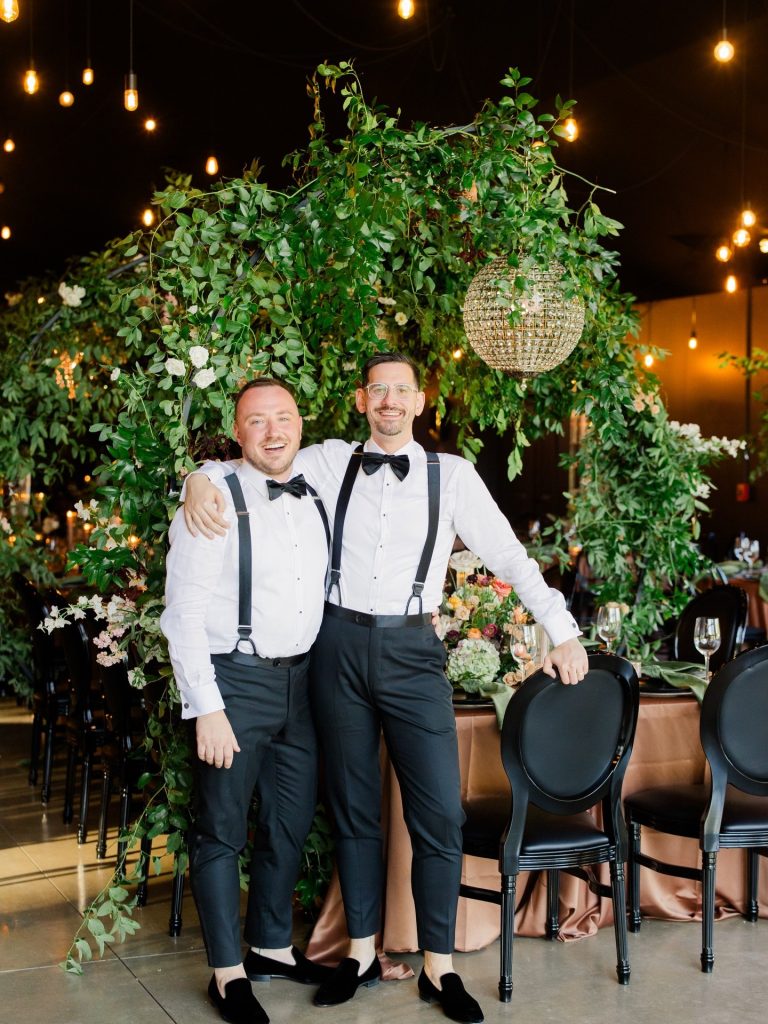 Stephen and Austin embrace in formal attire beneath dramatic hanging greenery and Edison bulb lighting at their reception