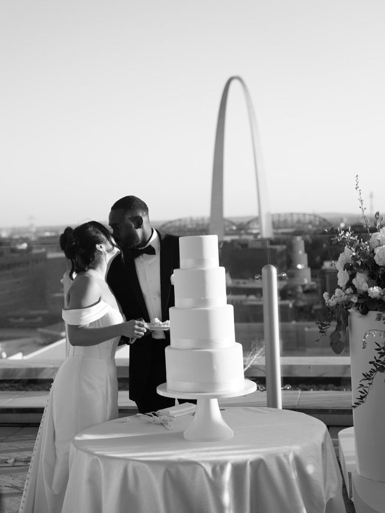 Katie and Anthony kiss behind their four-tier white wedding cake on rooftop terrace overlooking Gateway Arch