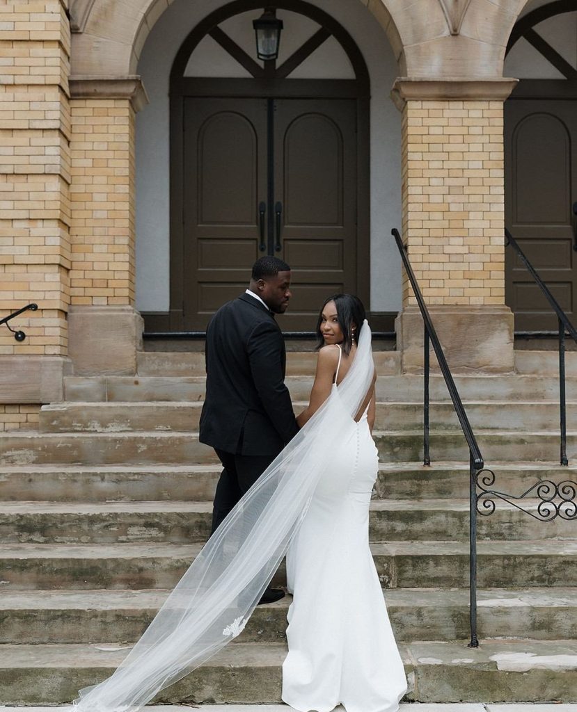 Newlyweds walk up stone steps with bride's cathedral-length veil flowing behind them outside historic building