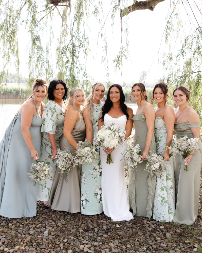 Hannah with bridesmaids in mixed sage green and floral dresses holding white and baby's breath bouquets under willow tree
