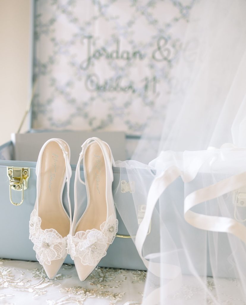 Bridal shoes with embroidered flowers displayed near vintage blue luggage and personalized handkerchief reading "Jordan & Clayton"