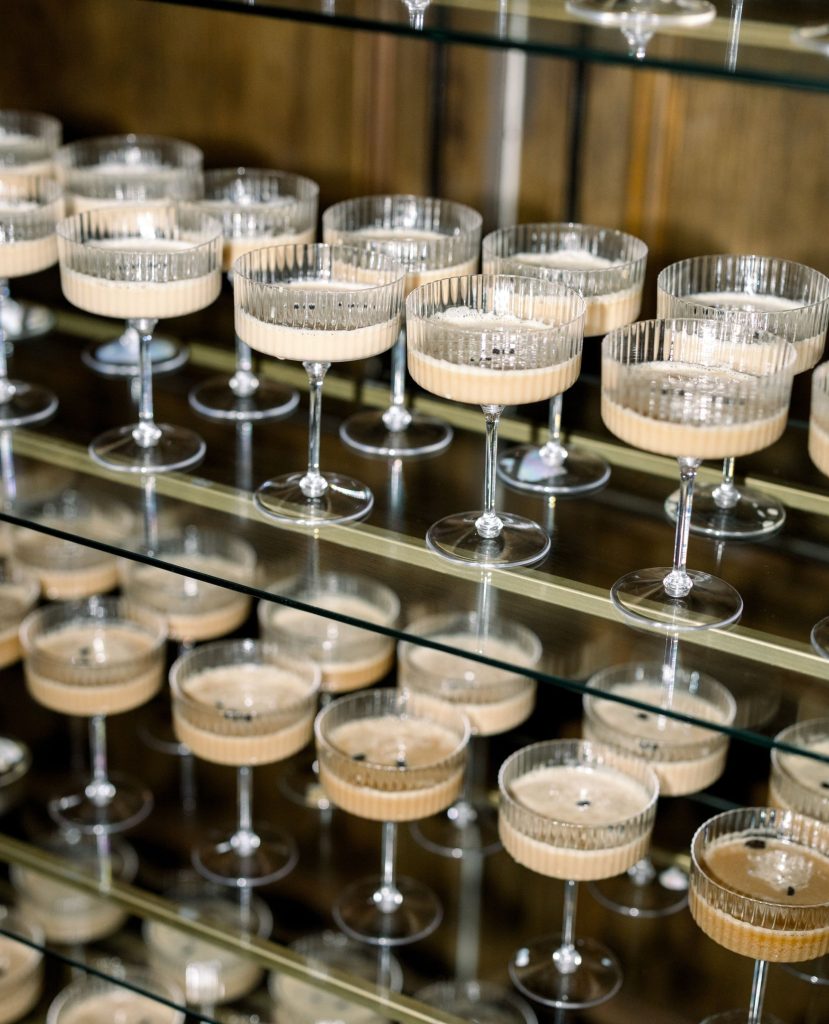 Champagne coupes arranged on glass shelves displaying cream-colored cocktails for wedding reception