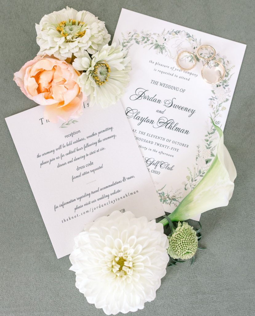 Wedding invitation suite with greenery border for Jordan and Clayton, displayed with wedding rings and white, peach, and cream flowers